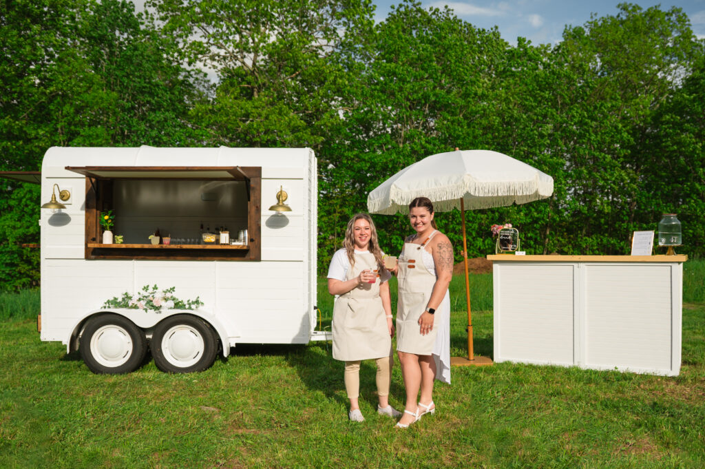 Luxury Mobile Bar & Balloon Service staff standing beside a mobile bar trailer with drinks in hand.